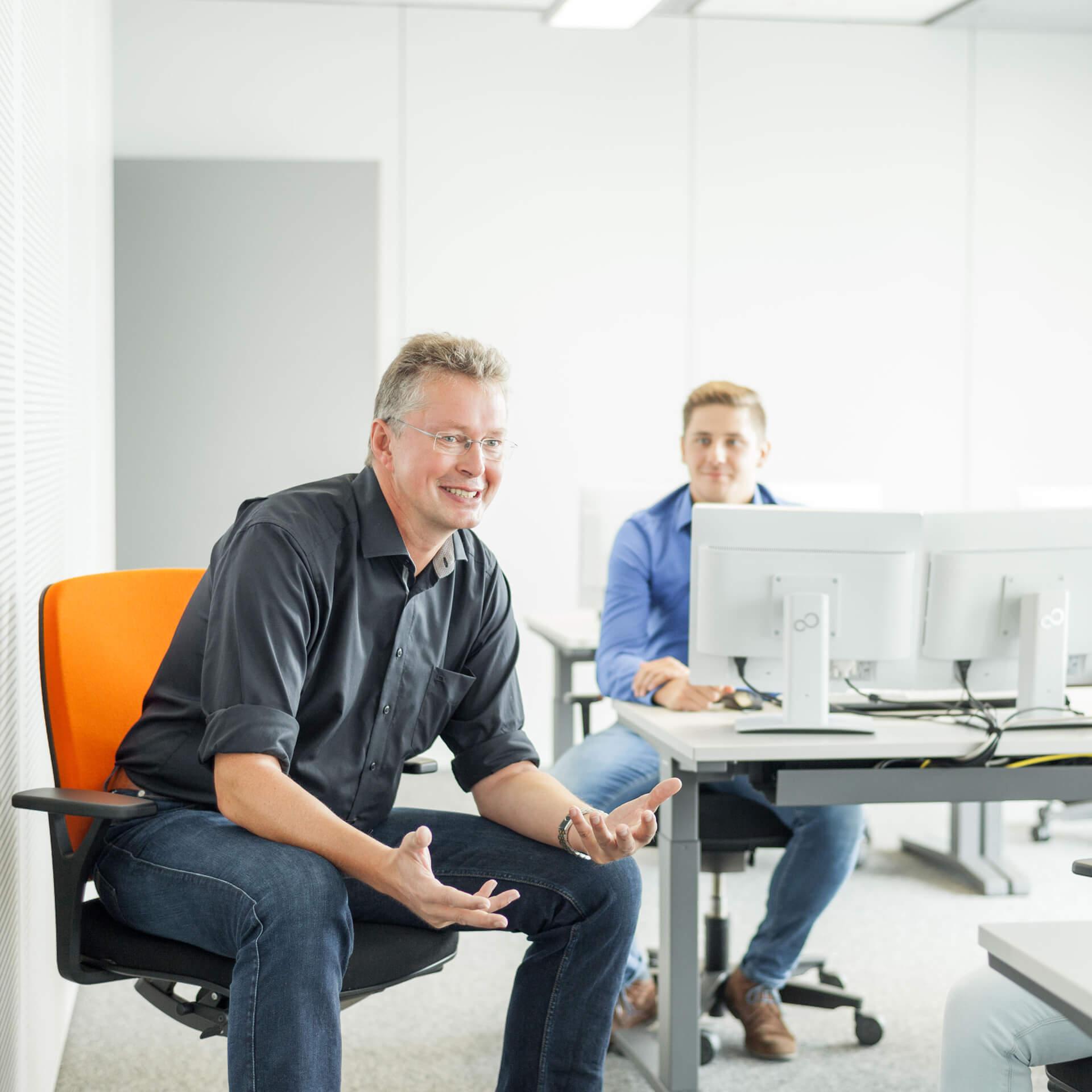 Mann in orangenem Bürostühl und zuhörender Mann am Bürotisch vor zwei Bildschirmen