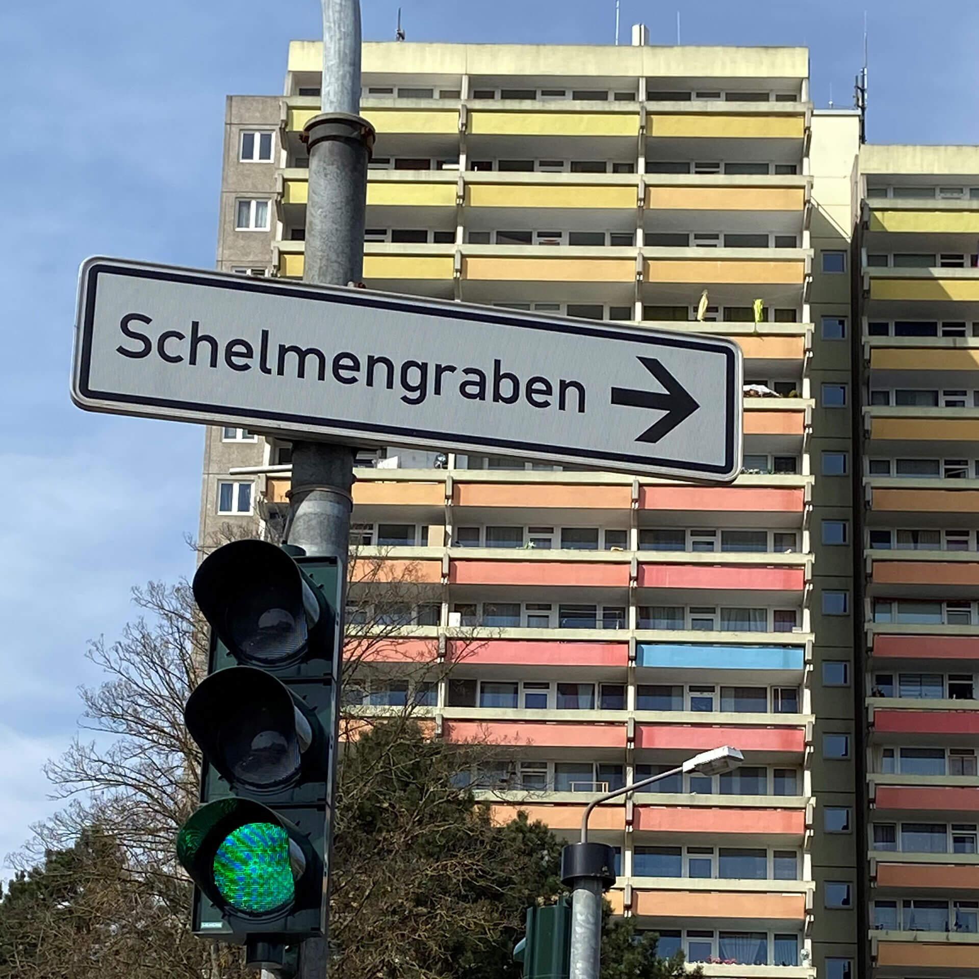 Grüne Ampel mit Straßenschild "Schelmengrabe" vor Hochhaus