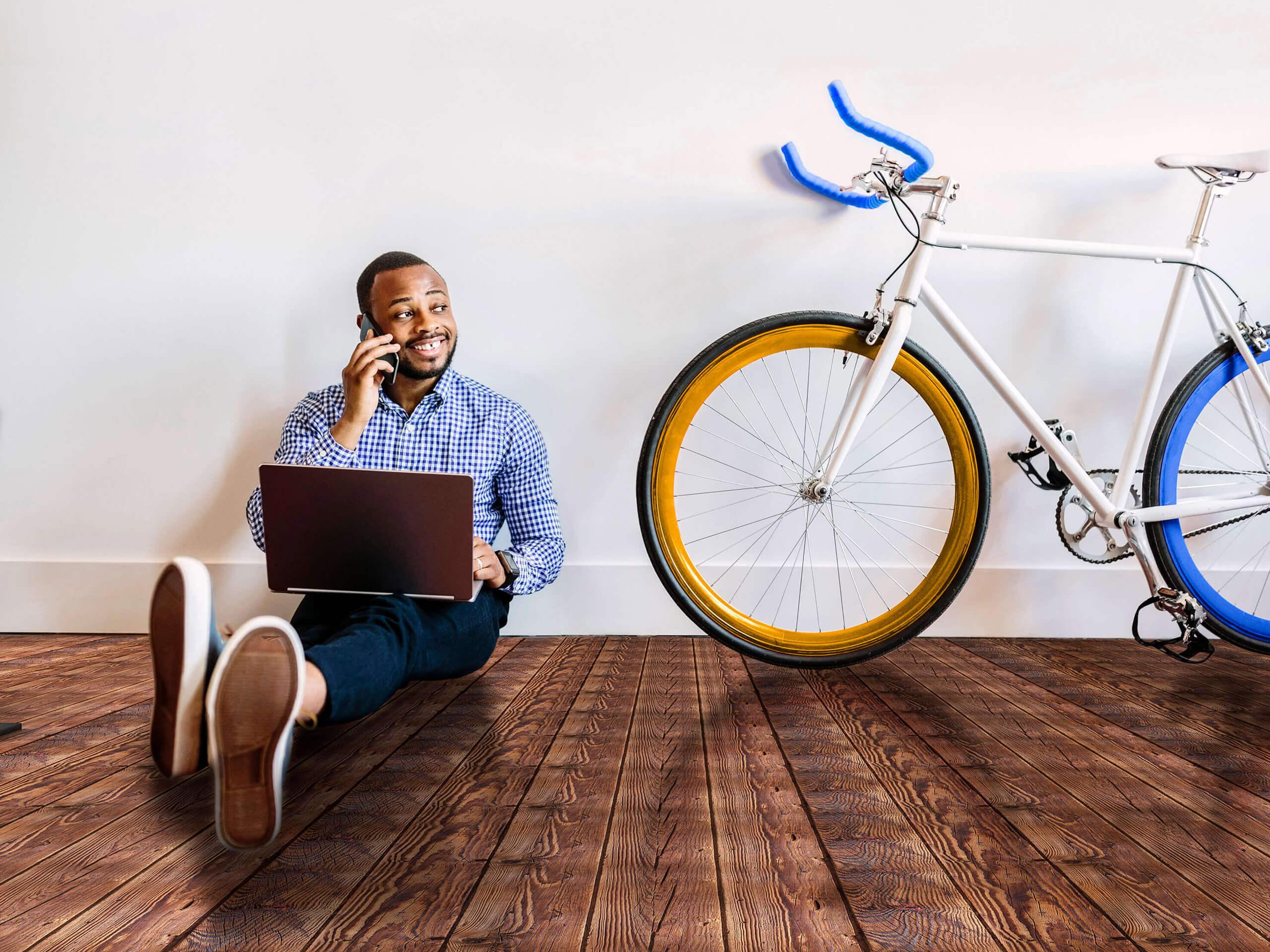 Sitzender Mann auf Holzboden mit Laptop und Handy sowie Rennrad vor weißer Wand